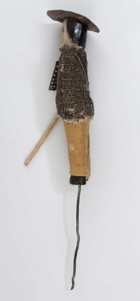 Figurka z szopki - blacharz. Kadłub drewniany na drucie. Twarz lekko ciosana. Rysy twarzy zaznaczony farbą. Blaszany kapelusz, szaro-czarna bluza, rude spodnie. Przez ramię przewieszona torba skórzana na pasku. Na piersi przymocowane narzędzia kuchenne z blachy. W prawym ręku drewniany kij.