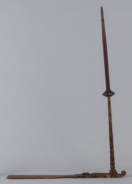 E/9283/ML - Przęślica drewniana wykonana z 3 części. Przysiadka w formie płaskiej deszczułki zwężająca się w jednym końcu i zdobiona snycerką: rozeta, motyw sercowaty, ząbki. Jej ostre zakończenie ma formę woluty/ślimaka. Przy węższym końcu osadzone pionowo ramię, toczone w pierścieniowate ozdoby. Na niego nakładany ruchomy krężel, również w toczony w pierścieniowate ozdoby. Przęślica powleczona brązowo-wiśniową farbą. Pierścieniowate elementy malowane na czarno, a na krężlu dodatkowo na srebrno.