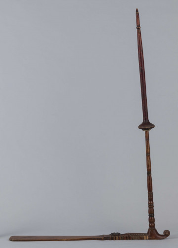 E/9283/ML - Przęślica drewniana wykonana z 3 części. Przysiadka w formie płaskiej deszczułki zwężająca się w jednym końcu i zdobiona snycerką: rozeta, motyw sercowaty, ząbki. Jej ostre zakończenie ma formę woluty/ślimaka. Przy węższym końcu osadzone pionowo ramię, toczone w pierścieniowate ozdoby. Na niego nakładany ruchomy krężel, również w toczony w pierścieniowate ozdoby. Przęślica powleczona brązowo-wiśniową farbą. Pierścieniowate elementy malowane na czarno, a na krężlu dodatkowo na srebrno.