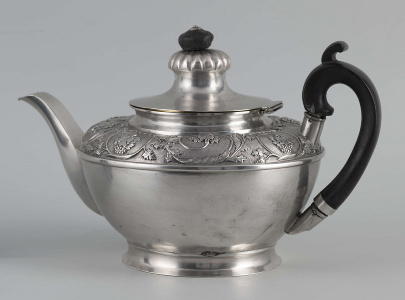 S/Mt/542/ML - Korpus w kształcie spłaszczonej kuli, na niskiej stopie, wylew w formie dzióbka, ucho drewniane malowane na czarno, osadzone w dwóch tulejkach, pokrywka na zawiasie, płaska, w części środkowej wypiętrzona i ozdobiona puklowaną rozetą.Górna część brzuśca ozdobiona sztancowaną taśmą z wzorem spiralnie zwinietej wici roślinnej z kwiatami, uchwyt przykrywki zakończony drewnianą rozetką.  
