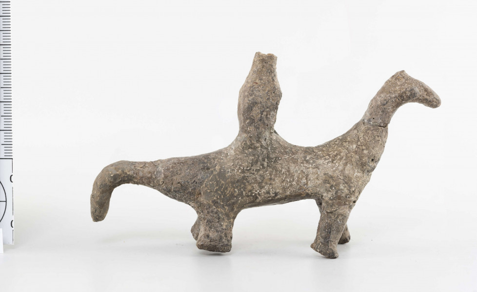 Figurka gliniana jeźdźca na koniu wykonana z wałka glinianego, szyjka wyciagnięta do góry, zwężona, zakończona spłaszczoną głową. Ogon mocno ścieniony, załamany do dołu. Kończyny walcowate, doklejone do korpusu. Postać siedząca na grzbiecie wykonana schematycznie. Surowiec - glina z domieszką piasku. Barwa brunatna. Stan zachowania dobry - drobne ubytki. Zabytek sklejany, uzupełniany gipsem.
Wymiary dodatkowe - 1,9 cm (grub. korpusu); 3 cm (wys. postaci); 1,4 cm (grub. postaci)