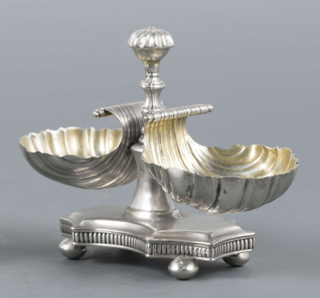 S/Mt/494/ML - Solniczka składa się z dwóch czarek w formie muszli połączonych z pionowym, profilowanym uchwytem o zwieńczniu w formie makówki ustawionym na wydłużonej ośmiobocznej podstawie o wklęsłych bokach. Krawędź podstawy fasetowana, boki karbowane, cztery nóżki w kształcie spłaszczonych kulek. Wnetrze muszli złocone.
Na dnie podstawy sygnatura kursywą w owalnych ramkach:
- J.Gałecki
- w Lublinie
- punca owalna z przedstawieniem dłoni (sygnatura pomocnicza)
- wężyk probierczy
- znak probierni warszawskiej z próbą 