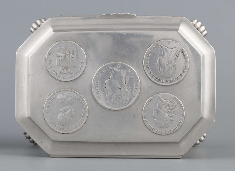 S/Mt/364/ML - Cukiernica skrzynkowa prostokątna ze ściętymi narożami, na czterech nóżkach w formie uskrzydlonych lwich łap, z wieczkiem o wklęsło profilowanym brzegu. Górny i dolny brzeg obiega  moletowana taśma z motywem neogotyckich trójliści, szyld zamka ma formę różanego wieńca pod skrzydatą główką..W ścianki i wieczko wmontowanych jest 15 monet srebrnych:
1 ort Zygmunta III
1 talar pruski, 1818 r.
1 pięciofrankówka, 1811 r.
1 moneta austriacka, 1797 r.
1 moneta saska, 
2 monety polskie St.Augusta Poniatowskiego
1 dwózłotówka Królestwa Polskiego, 1825 r.
1 pięciozłotówka, 1831 r.
6 złotówek, 1831 r.
Na dnie sygnatura wklęsła kursywą:
- K. Rotkiel w Lublinie
i punca z oznaczeniem próby 