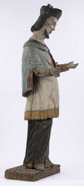 E/8422/ML - Rzeźba drewniana, pełna, polichromowana, przedstawiająca św. Jana Nepomucena. Zakomponowana frontalnie, z rękoma zgiętymi w łokciu i lekko rozłożonymi. Twarz owalna, pomalowana białą farbą. Małą broda i krótkie czarne wąsy. Nos krótki, prosty. Oczy owalne podkreślone czarnym konturem. Brwi łukowate, wąskie, czarne. Włosy czarne, ułożone w pukle. Na głowie czarny biret. Długa czarna sutanna, biała komża ozdobiona u dołu złotą koronką. Na piersiach krzyż, zawieszony na łańcuchu. Na komżę narzucona jasnoniebieska peleryna. Stopy lekko rozstawione. Obuty w sandały o jednym pasku. 

Konserwacja: 1990 r.