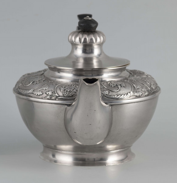 S/Mt/542/ML - Korpus w kształcie spłaszczonej kuli, na niskiej stopie, wylew w formie dzióbka, ucho drewniane malowane na czarno, osadzone w dwóch tulejkach, pokrywka na zawiasie, płaska, w części środkowej wypiętrzona i ozdobiona puklowaną rozetą.Górna część brzuśca ozdobiona sztancowaną taśmą z wzorem spiralnie zwinietej wici roślinnej z kwiatami, uchwyt przykrywki zakończony drewnianą rozetką.  
