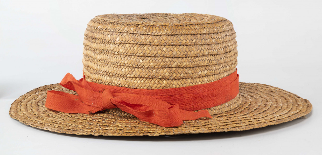 Kapelusz ze słomy owsianej o prostym denku i rondzie płaskim. Upleciony techniką warkoczową. Warkocze bez ząbków, zszyte cienkimi szarymi nićmi. Denko otoczone czerwoną wąską wstążką, zakończone kokardą. 