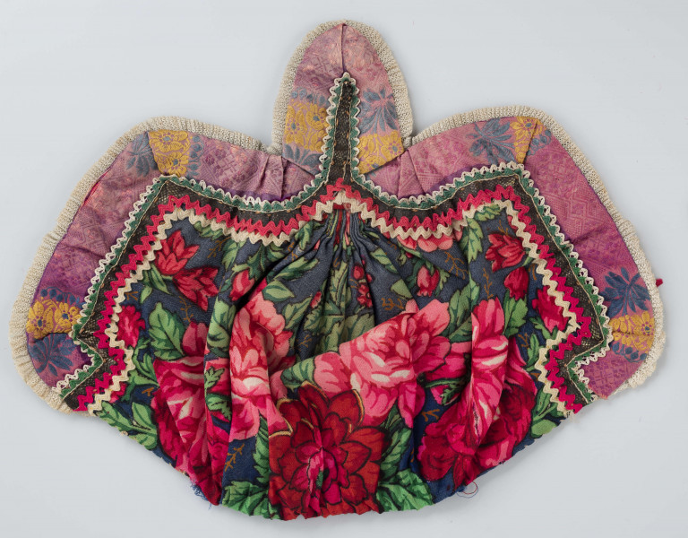 Czepek mężatki na płóciennej podszewce z granatowej cienkiej wełenki w amarantowe róże i jasnozielone listki. Marszczony na środku przodu i tyłu. Przód ozdobionymi kremowymi i amarantowymi ząbkami, srebrną metaliczną wstążką oraz szeroką atłasową wstążką w kolorze fioletowym w granatowe i żółte kwiaty. Brzeg wykończony białą tiulową, drobno plisowaną kryzą. Przód wycięty w duży ząb w kształt płatka kwiatu.