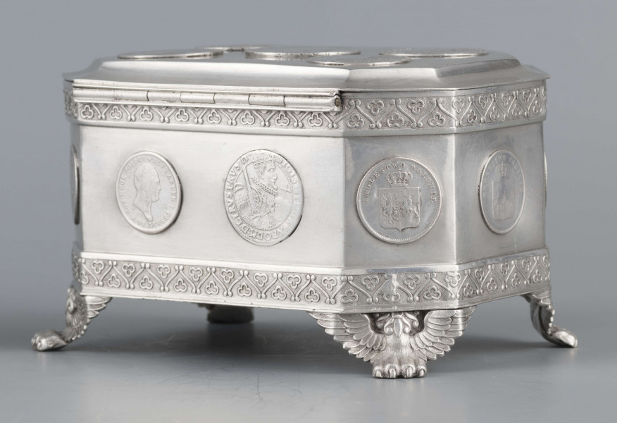 S/Mt/364/ML - Cukiernica skrzynkowa prostokątna ze ściętymi narożami, na czterech nóżkach w formie uskrzydlonych lwich łap, z wieczkiem o wklęsło profilowanym brzegu. Górny i dolny brzeg obiega  moletowana taśma z motywem neogotyckich trójliści, szyld zamka ma formę różanego wieńca pod skrzydatą główką..W ścianki i wieczko wmontowanych jest 15 monet srebrnych:
1 ort Zygmunta III
1 talar pruski, 1818 r.
1 pięciofrankówka, 1811 r.
1 moneta austriacka, 1797 r.
1 moneta saska, 
2 monety polskie St.Augusta Poniatowskiego
1 dwózłotówka Królestwa Polskiego, 1825 r.
1 pięciozłotówka, 1831 r.
6 złotówek, 1831 r.
Na dnie sygnatura wklęsła kursywą:
- K. Rotkiel w Lublinie
i punca z oznaczeniem próby 