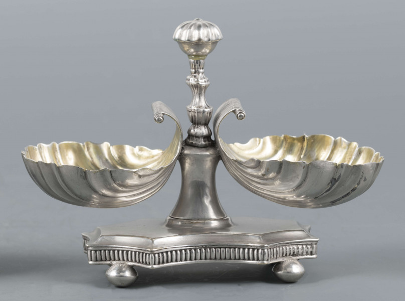 S/Mt/494/ML - Solniczka składa się z dwóch czarek w formie muszli połączonych z pionowym, profilowanym uchwytem o zwieńczniu w formie makówki ustawionym na wydłużonej ośmiobocznej podstawie o wklęsłych bokach. Krawędź podstawy fasetowana, boki karbowane, cztery nóżki w kształcie spłaszczonych kulek. Wnetrze muszli złocone.
Na dnie podstawy sygnatura kursywą w owalnych ramkach:
- J.Gałecki
- w Lublinie
- punca owalna z przedstawieniem dłoni (sygnatura pomocnicza)
- wężyk probierczy
- znak probierni warszawskiej z próbą 