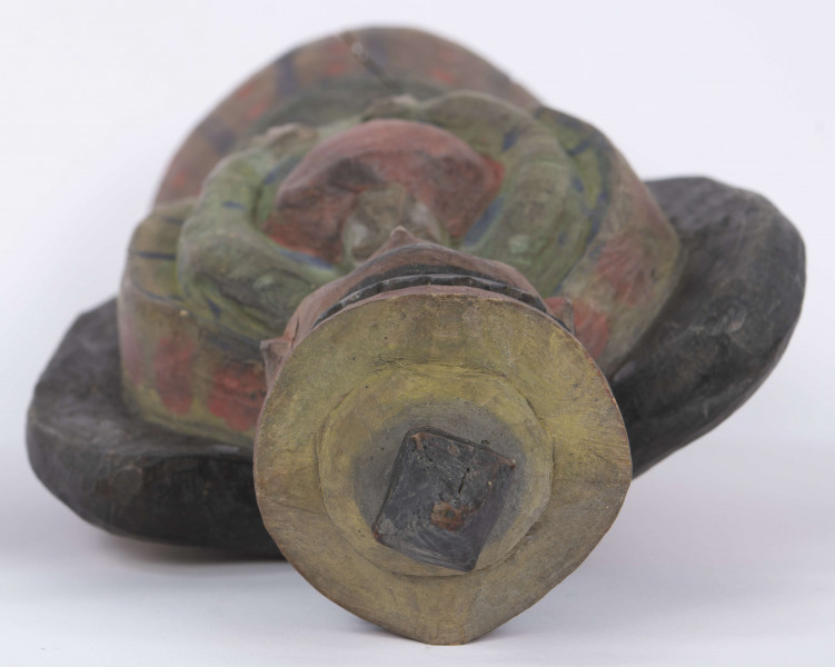 E/8100/ML - Rzeźba pełna wykonana z jednego kawałka drewna lipowego, polichromowana. Postać w hieratycznej postaci, w układzie frontalnym. Ręce wsparte na biodrach. Postać stoi na stożkowatym cokole. Na głowie korona z dwóch oddzielnych kawałków drewna, złączonych ozdobnym czopem drewnianym. Włosy czarne, ręce i twarz różowa, korona żółta z czerwonym obrzeżem, zwieńczenie czarne. Płaszcz stylizowany, przypominający skrzydła lub ornat, czarny. Szaty wewnętrzne żółte z czerwonymi plamkami. Wewnątrz długie, zielone z niebieskimi plamami. Buty czarne. Na piersiach duże otwarte serce zwieńczone czarnym krzyżem. Cokół w naturalnym kolorze drewna, ozdobiony pionowymi niebieskimi paskami i czerwonymi kropkami. Wym. cokołu: 13 x 10,5 cm. Sygnatura na spodzie (pisana): Jan Kaproń.