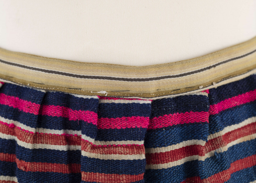 E/7351/ML - Zapaska wełniana w granatowe, czerwone, białe i amarantowe paski biegnące w poprzek. Zszyta z dwóch części – pionowy szew na przodzie. Dołem ozdobiona szerokim szlakiem dekoracyjnym, złożonym z pasków: zielonych, żółtych i amarantowych. W pasie marszczona i ujęta w oszewkę z innego materiału.