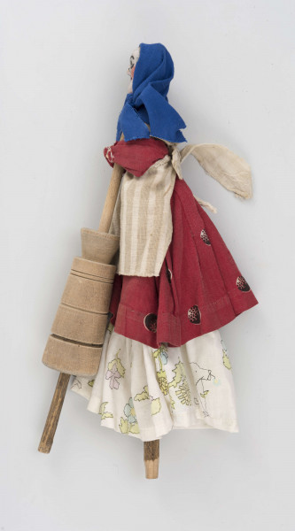 Kukiełka z szopki przedstawiająca czarownicę robiącą masło w maselnicy. Postać ubrana w jasną długą spódnicę, czerwony w koła kaftan i fartuch w szare pionowe pasy na jasnym tle. Na głowie ma granatową jedwabną chustkę wiązaną pod brodą. Twarz słabo modelowana, nos wystający, oczy namalowane farbą. Maselnica toczona, przyczepiona drutem do kukiełki.