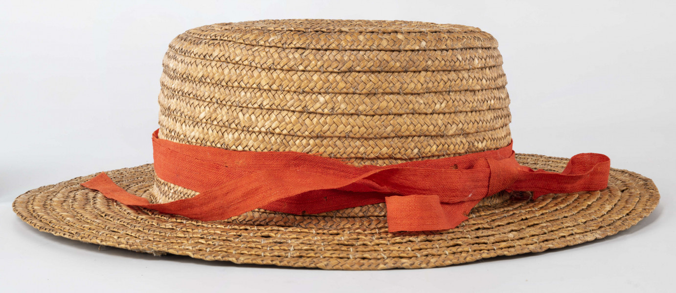 Kapelusz ze słomy owsianej o prostym denku i rondzie płaskim. Upleciony techniką warkoczową. Warkocze bez ząbków, zszyte cienkimi szarymi nićmi. Denko otoczone czerwoną wąską wstążką, zakończone kokardą. 