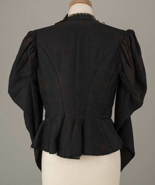 E/8038/ML - Kaftan kobiecy zszyty z 10 kawałków czarnej wełenki. Składa się z 2 części przodu, pleców i rękawów. Wycięcie wokół szyi wykończone stójką szer. 3,5 cm. Przód zapinany z boku na haftki. Rękawy w góry bufiaste, suto marszczone, zwężające się. Przód ułożony w zakładki. Stójka i przód ozdobiony żółtym sznureczkiem, czarną sieczką i białymi szklanymi koralikami. Szyty na maszynie.