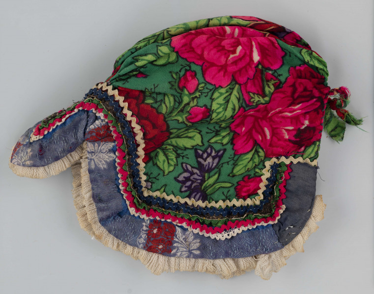 E/8141/ML - Czepek mężatki na płóciennej podszewce z zielonej cienkiej wełenki w amarantowe róże, fioletowe sasanki i jasnozielone listki. Marszczony na środku przodu i tyłu. Przód ozdobionymi kremowymi i granatowymi ząbkami, srebrną metaliczną wstążką oraz szeroką atłasową wstążką w kolorze granatowym w białe i czerwone kwiaty. Brzeg wykończony białą tiulową, drobno plisowaną kryzą. Przód wycięty w duży ząb w kształt płatka kwiatu.