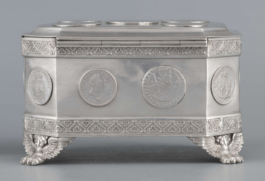 S/Mt/364/ML - Cukiernica skrzynkowa prostokątna ze ściętymi narożami, na czterech nóżkach w formie uskrzydlonych lwich łap, z wieczkiem o wklęsło profilowanym brzegu. Górny i dolny brzeg obiega  moletowana taśma z motywem neogotyckich trójliści, szyld zamka ma formę różanego wieńca pod skrzydatą główką..W ścianki i wieczko wmontowanych jest 15 monet srebrnych:
1 ort Zygmunta III
1 talar pruski, 1818 r.
1 pięciofrankówka, 1811 r.
1 moneta austriacka, 1797 r.
1 moneta saska, 
2 monety polskie St.Augusta Poniatowskiego
1 dwózłotówka Królestwa Polskiego, 1825 r.
1 pięciozłotówka, 1831 r.
6 złotówek, 1831 r.
Na dnie sygnatura wklęsła kursywą:
- K. Rotkiel w Lublinie
i punca z oznaczeniem próby 