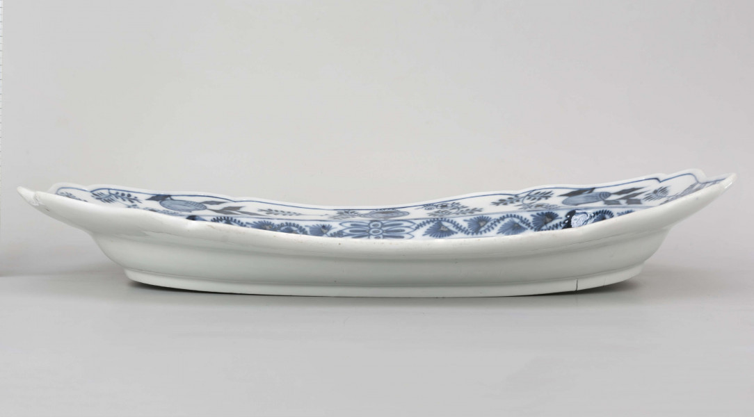 Półmisek owalny, kołnierz gładki o brzegu wykrojonym faliscie. Malowany podszkliwnie kobaltem we wzór tzw. cebulowy, na dnie wieksza kompozycja ze stylizowanych gałązek z kwiatami. Dno wzmocnione od spodu esowatym pasmem z masy porcelanowej.
 