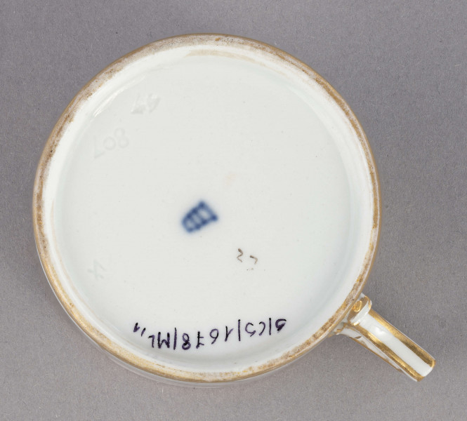 Filiżanka i spodek malowane w piórka. Filiżanka cylindryczna, ucho łamane z listkami w osadzeniach. Brzeg złocony obustronnie, złotem  konturowane ucho, złote kontury dzielą powierzchnię na ukośne pasy na przemian białe i jasnoszare. W nich malowane odpowiednio złote i czarne pióra.