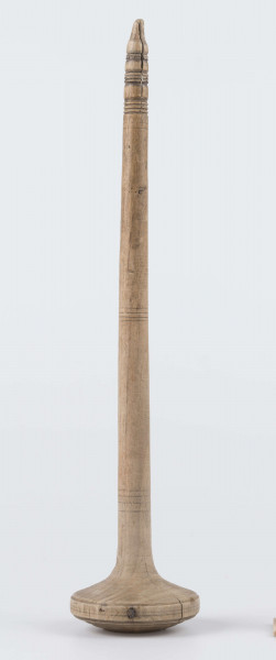 E/8019/ML - Przęślica krężołkowa z drewna brzozowego składa się z cienkiej deski – przysiadki, w której przy jednym końcu osadzona jest pionowa beleczka zw. piórem, z nasadzonym od góry ruchomym krężlem w kształcie wydłużonego stożka. Deseczka przysiadki o kształcie prostokątnym lekko zwężającym, od ok. połowy swej długości w kierunku pióra, i zakończonym głową zwierzęcą – psa. Pióro i krężel ozdobnie toczone. Krężel ma kształt stożka z rozszerzoną podstawą, w której znajduje się stożkowaty otwór, do osadzenia krężla na piórze. Dekoracja snycerska ryta znajduje się na zwężającej części przysiadki. Część zdobiona oddzielona małym stopniem. Grubość przysiadki w części zdobionej 2x większa od pozostałej części. Dekoracja snycerska tworzy ornament roślinny w postaci trzech ulistnionych drobno gałązek. Na zakończeniu ornamentu znajdują się ryte liście oraz inicjały „K.F.”. Głowa psa zdobiona drobnymi nacięcia. Zakończenie przysiadki wycięte faliście.