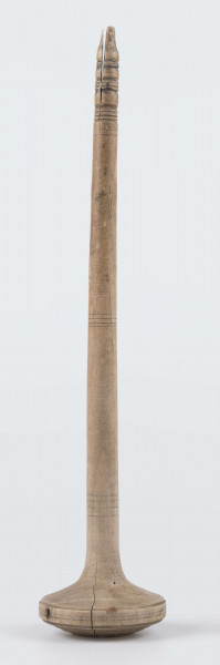 E/8019/ML - Przęślica krężołkowa z drewna brzozowego składa się z cienkiej deski – przysiadki, w której przy jednym końcu osadzona jest pionowa beleczka zw. piórem, z nasadzonym od góry ruchomym krężlem w kształcie wydłużonego stożka. Deseczka przysiadki o kształcie prostokątnym lekko zwężającym, od ok. połowy swej długości w kierunku pióra, i zakończonym głową zwierzęcą – psa. Pióro i krężel ozdobnie toczone. Krężel ma kształt stożka z rozszerzoną podstawą, w której znajduje się stożkowaty otwór, do osadzenia krężla na piórze. Dekoracja snycerska ryta znajduje się na zwężającej części przysiadki. Część zdobiona oddzielona małym stopniem. Grubość przysiadki w części zdobionej 2x większa od pozostałej części. Dekoracja snycerska tworzy ornament roślinny w postaci trzech ulistnionych drobno gałązek. Na zakończeniu ornamentu znajdują się ryte liście oraz inicjały „K.F.”. Głowa psa zdobiona drobnymi nacięcia. Zakończenie przysiadki wycięte faliście.