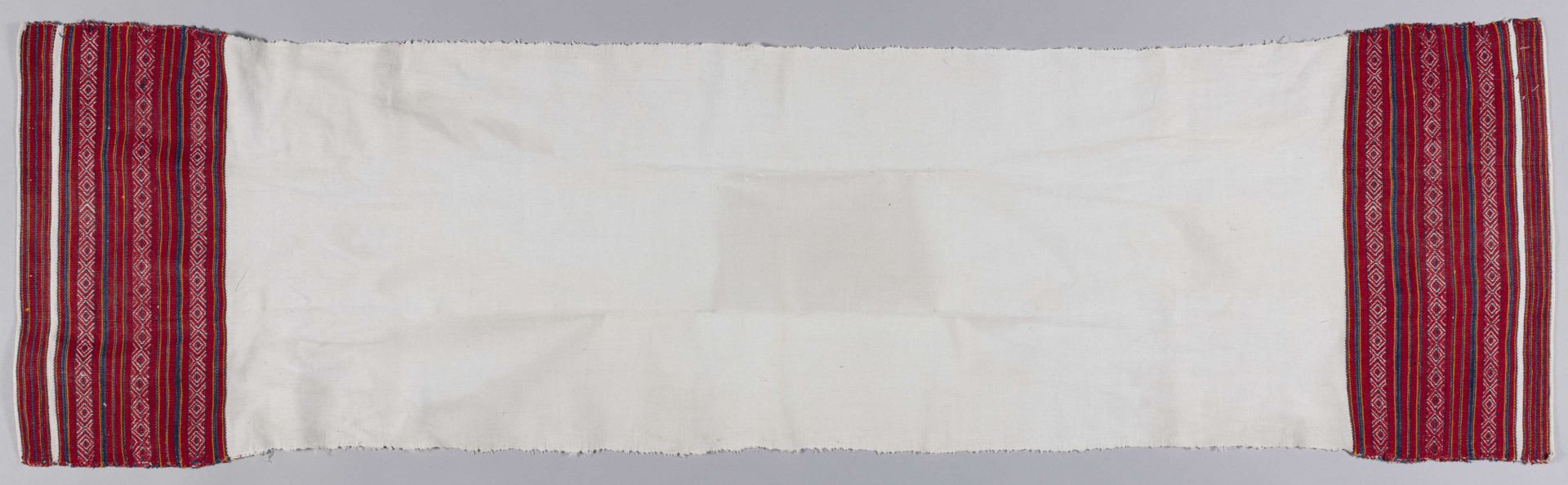 E/4943/ML - Ręcznik lniany, z haftem na obu końcach. Haft tkany na warsztacie. Wzór tworzą pasy w kolorach: czerwonym, pomarańczowym i granatowym oraz szlak złożony z rombów i serduszek. Szerokość kolorowego szlaku - 16 cm.