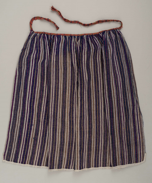 E/5997/ML - Fartuszek uszyty z jednego kawałka materiału - tkaniny samodziałowej bawełniano-wełnianej, tkanej splotem rypsowym w drobne paseczki w kolorze: białym, czerwonym, granatowym i bordo. Suto marszczony w oszewkę przechodzącą w troczki. Oszewka z materiału lnianego w kratkę czerwono-zielono-granatową. Dół obszyty tasiemką z białego bawełnianego płótna.