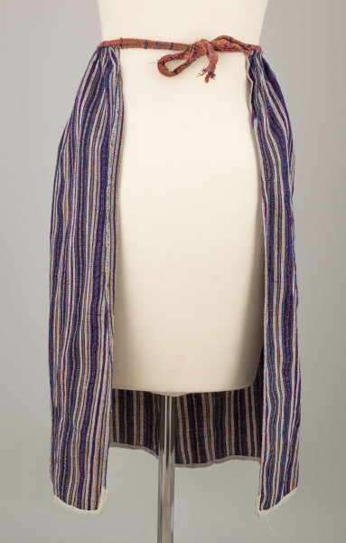 E/5997/ML - Fartuszek uszyty z jednego kawałka materiału - tkaniny samodziałowej bawełniano-wełnianej, tkanej splotem rypsowym w drobne paseczki w kolorze: białym, czerwonym, granatowym i bordo. Suto marszczony w oszewkę przechodzącą w troczki. Oszewka z materiału lnianego w kratkę czerwono-zielono-granatową. Dół obszyty tasiemką z białego bawełnianego płótna.