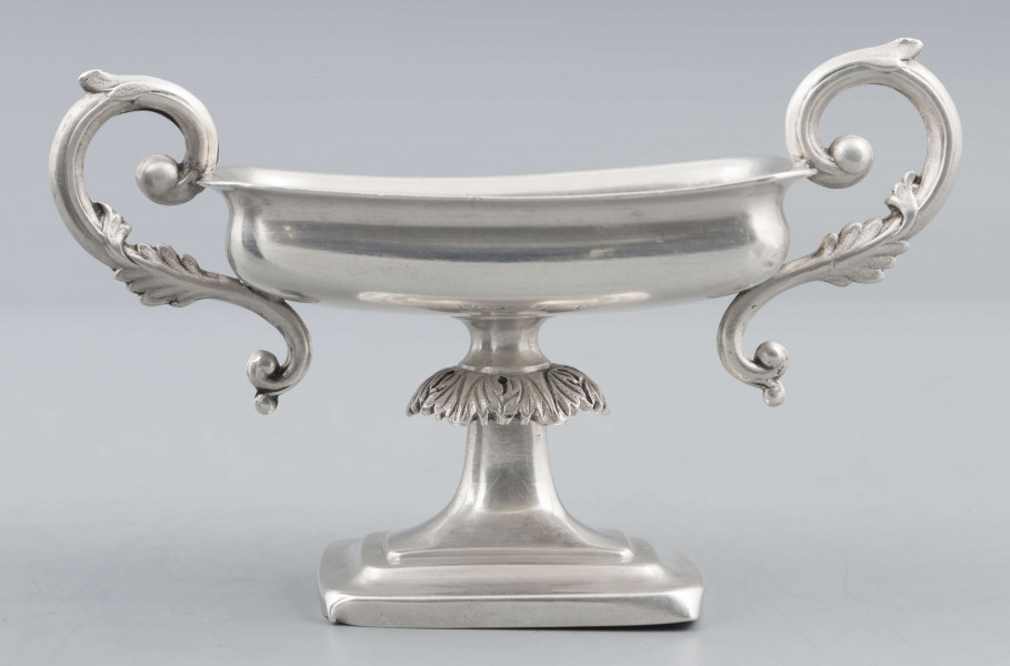 S/Mt/57/ML - Stopa prostokątna, wysklepiona, przechodząca w trzon z owalnym kapturkiem sztancowanym we wzór akantu. Czarka prostokatna o zaokrąglonych narożach i wywinietym brzegu, z dwoma uchami na krótszych bokach. Ucha wolutowe zdobione listkami. 