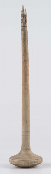 E/8019/ML - Przęślica krężołkowa z drewna brzozowego składa się z cienkiej deski – przysiadki, w której przy jednym końcu osadzona jest pionowa beleczka zw. piórem, z nasadzonym od góry ruchomym krężlem w kształcie wydłużonego stożka. Deseczka przysiadki o kształcie prostokątnym lekko zwężającym, od ok. połowy swej długości w kierunku pióra, i zakończonym głową zwierzęcą – psa. Pióro i krężel ozdobnie toczone. Krężel ma kształt stożka z rozszerzoną podstawą, w której znajduje się stożkowaty otwór, do osadzenia krężla na piórze. Dekoracja snycerska ryta znajduje się na zwężającej części przysiadki. Część zdobiona oddzielona małym stopniem. Grubość przysiadki w części zdobionej 2x większa od pozostałej części. Dekoracja snycerska tworzy ornament roślinny w postaci trzech ulistnionych drobno gałązek. Na zakończeniu ornamentu znajdują się ryte liście oraz inicjały „K.F.”. Głowa psa zdobiona drobnymi nacięcia. Zakończenie przysiadki wycięte faliście.