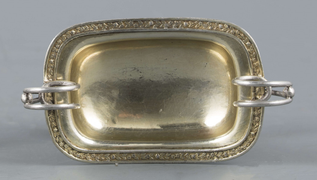 S/Mt/359/ML - Solniczka w kształcie prostokątnej łódki z dwoma wolutowymi uchami z okrągłego drutu, umieszczonej na figurce gryfa, z  podstawą prostokątną o wklęsłych narożach i czterech nóżkach w formie spłaszczonych kulek. Brzeg łódki wywinięty, wnętrze złocone. Brzegi łódki i boki podstawy obiega moletowana taśma z ornamentem roślinnym.