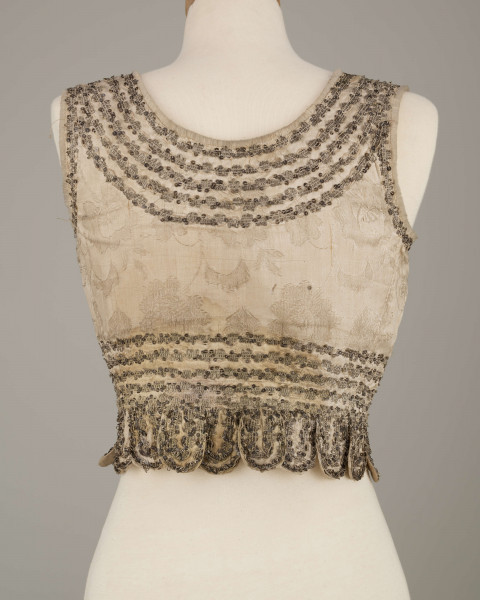 E/6235/ML - Gorset ślubny z białego wzorzystego atłasu. Składa się z 3 części: pleców i dwóch przodów. W pasie doszyte 17 owalnych klapek, zw. taszki. Wycięcie wokół szyi , przodów i rękawów wykończone białą wełnianą tasiemką, zw. szczotka. Całość zdobiona złotym szychem i srebrnymi cekinami. Podszyty białą bawełnianą podszewką.  