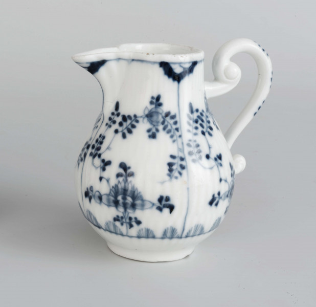 Dzbanek gruszkowaty na niskiej pierścieniowatej podstawie, ucho wolutowe, wylew trókątny, powierzchnia drobno kanelowana. Malowany podszkliwnie błękitem kobaltowym we wzór suchych kwiatów (kocanka), poniżej brzegu górnego na zewnatrz i wewnątrz malowana cienka obwódka, na dnie brzegu uszka gałązka. 
