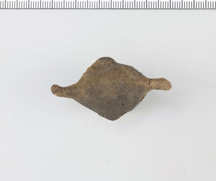Figurka zoomorficzna w kształcie ptaszka, wykonana schematycznie. Nóżka walcowata. Główka, ogon oraz skrzydełka ledwie zaznaczone. Powierzchnia gładzona niestarannie. Sklejone, uzupełnione gipsem. Surowiec - glina z domieszką drobnoziarnistą. Barwa Szarobrunatna. Wypał dobry.
Wymiary dodatkowe: 1,3 cm (śr. stopki), 2,4 cm (rozstaw skrzydełek)