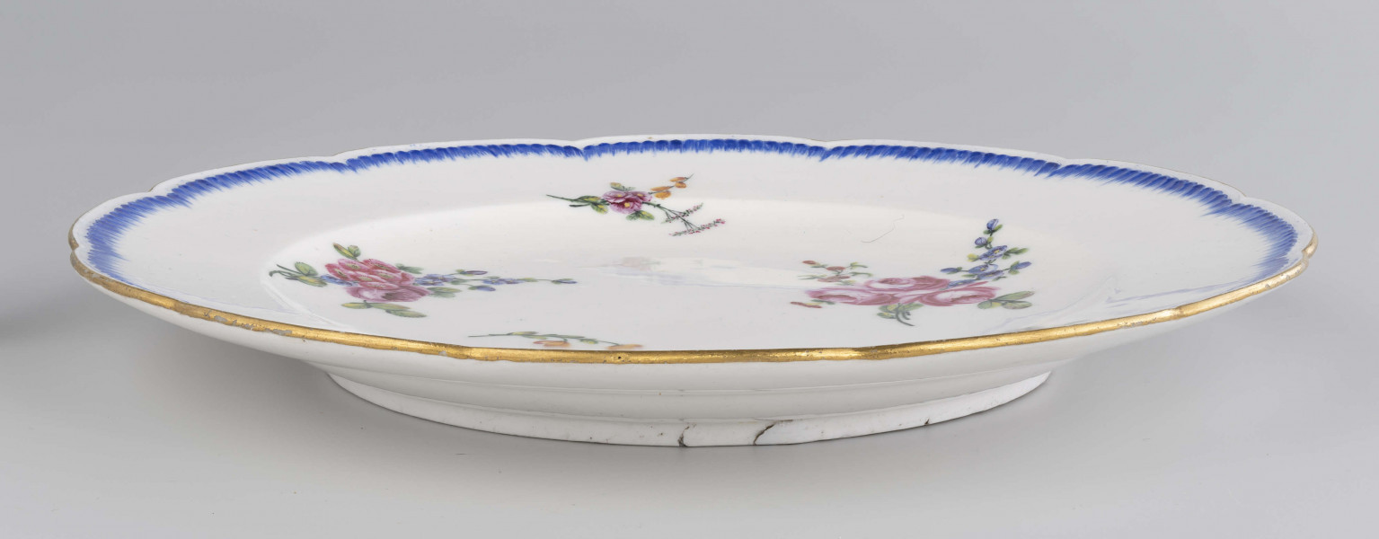 Talerz płytki o falistym wykroju brzegu, przy krawędzi plastyczne kanelowanie podkreślone błękitnym paskiem. Dekoracja malowana wielobarwnie: na całej powierzchni swobodnie rozrzucone różnej wielkości bukiety i gałązki kwiatowe.