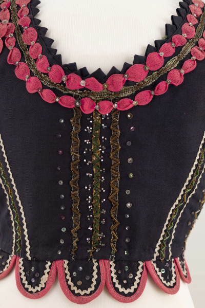 E/7358/ML - Gorset zszyty z 3 kawałków granatowej wełenki, podszyty brązową bawełnianą podszewką. Z przodu rozcięcie usztywnione fiszbinami, zaopatrzone w 10 haftek drucianych do przewlekania wstążeczki. Od pasa gorsetu dookoła opadają klapki. Cały gorset bogato zdobiony amarantowymi tasiemkami, żółtym i srebrnym szychem, białą krepiną, barwnymi koralikami i cekinami. Przód gorsetu, brzegi wykroju i pachy ozdobione dodatkowo granatowymi ząbkami. 