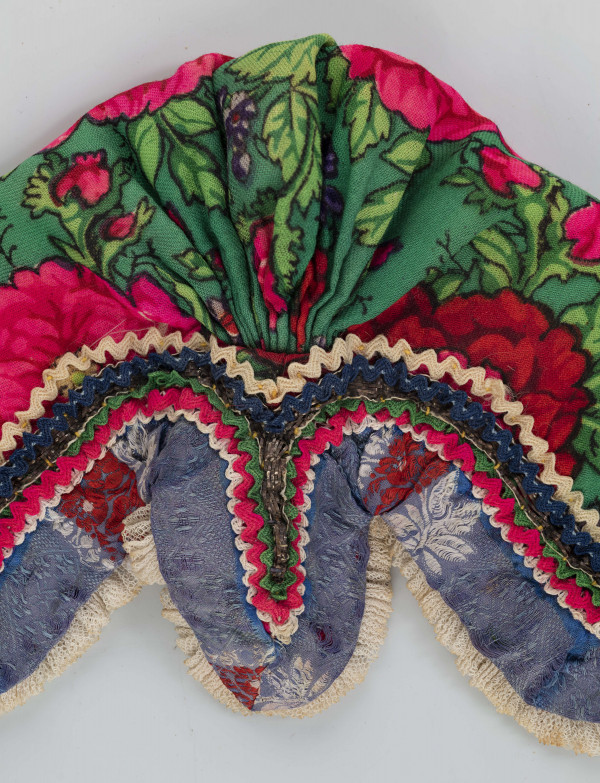 E/8141/ML - Czepek mężatki na płóciennej podszewce z zielonej cienkiej wełenki w amarantowe róże, fioletowe sasanki i jasnozielone listki. Marszczony na środku przodu i tyłu. Przód ozdobionymi kremowymi i granatowymi ząbkami, srebrną metaliczną wstążką oraz szeroką atłasową wstążką w kolorze granatowym w białe i czerwone kwiaty. Brzeg wykończony białą tiulową, drobno plisowaną kryzą. Przód wycięty w duży ząb w kształt płatka kwiatu.