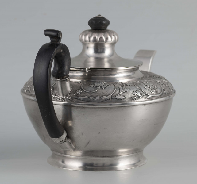 S/Mt/542/ML - Korpus w kształcie spłaszczonej kuli, na niskiej stopie, wylew w formie dzióbka, ucho drewniane malowane na czarno, osadzone w dwóch tulejkach, pokrywka na zawiasie, płaska, w części środkowej wypiętrzona i ozdobiona puklowaną rozetą.Górna część brzuśca ozdobiona sztancowaną taśmą z wzorem spiralnie zwinietej wici roślinnej z kwiatami, uchwyt przykrywki zakończony drewnianą rozetką.  
