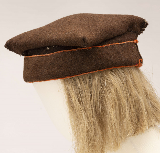 Czapka męska rogatywka szyta ręcznie z samodziałowego sukna w kolorze brązowym. Jeden pas stanowi otok., drugi tej samej szerokości jest wywinięty na wierzch i obszyty na dolnej i górnej krawędzi pomarańczowym wełnianym sznureczkiem. Górna cześć czapki wykonana z kwadratowego płata z wywiniętymi narożnikami, które po zszyciu i przyszyciu do otoka tworzą płaskie kwadratowe denko, o wym. 20 x 20 cm. Noszona do sukmany.