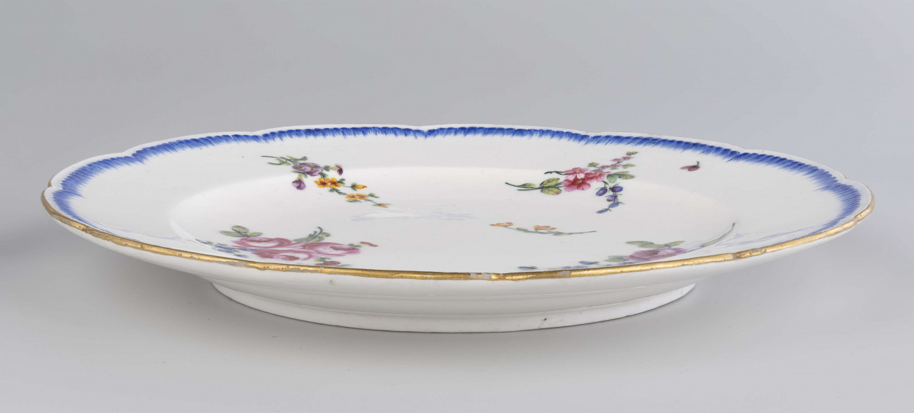 Talerz płytki o falistym wykroju brzegu, przy krawędzi plastyczne kanelowanie podkreślone błękitnym paskiem. Dekoracja malowana wielobarwnie: na całej powierzchni swobodnie rozrzucone różnej wielkości bukiety i gałązki kwiatowe.