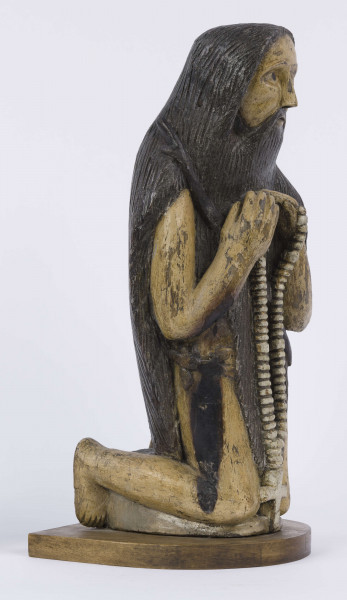 E/6353/ML - Figura drewniana, zakomponowana na postumencie, polichromowana. Postać klęcząca przedstawia św. Onufrego. Twarz płaska, trójkątna, głęboko osadzone oczy, czarne źrenice. Szerokie łuki brwiowe łączą się z wydatnym prostym nosem. Wystające kości jarzmowe. Małe usta otacza bujny zarost: broda i wąsy łączące się z bujnymi włosami, otaczającymi całą postać – na podobieństwo płaszcza. Ręce zgięte w łokciach, uniesione ku górze, łączą się końcami palców w geście modlitwy. W nieproporcjonalnie dużych dłoniach biały różaniec zakończony dużym krzyżem. Nogi masywne z dużymi płaskimi stopami. Prawą dłonią podtrzymuje sękaty kostur. W partii pasa przepasany powrozem z węzłami. 