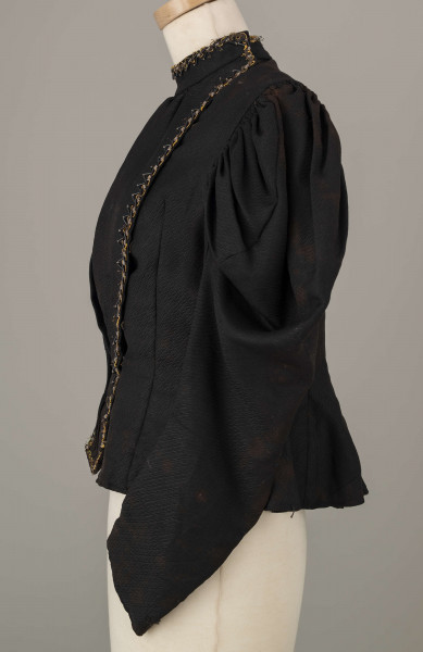 E/8038/ML - Kaftan kobiecy zszyty z 10 kawałków czarnej wełenki. Składa się z 2 części przodu, pleców i rękawów. Wycięcie wokół szyi wykończone stójką szer. 3,5 cm. Przód zapinany z boku na haftki. Rękawy w góry bufiaste, suto marszczone, zwężające się. Przód ułożony w zakładki. Stójka i przód ozdobiony żółtym sznureczkiem, czarną sieczką i białymi szklanymi koralikami. Szyty na maszynie.