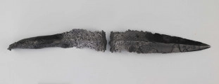Żelazny grot oszczepu z ostrym żeberkiem środkowym, zgięty rytualnie (złamany w miejscu zgięcia). Grot stanowi element wyposażenia ciałopalnego pochówku złożonego prawdopodobnie w popielnicy. Odkrycia dokonano w 1959 roku przypadkowo w czasie prac rolnych. Oprócz trzech grotów, pochówkowi wojownika towarzyszyły: żelazne umbo, dwie ostrogi, tok brązowy (prawdopodobnie okucie drzewca oszczepu) a także żelazny miecz z miedziana inkrustacją wyobrażającą rzymskich bogów. Pochowany na terenie obecnej wsi Podlodów wojownik należał do elity wojskowej przedstawicieli kultury przeworskiej.
