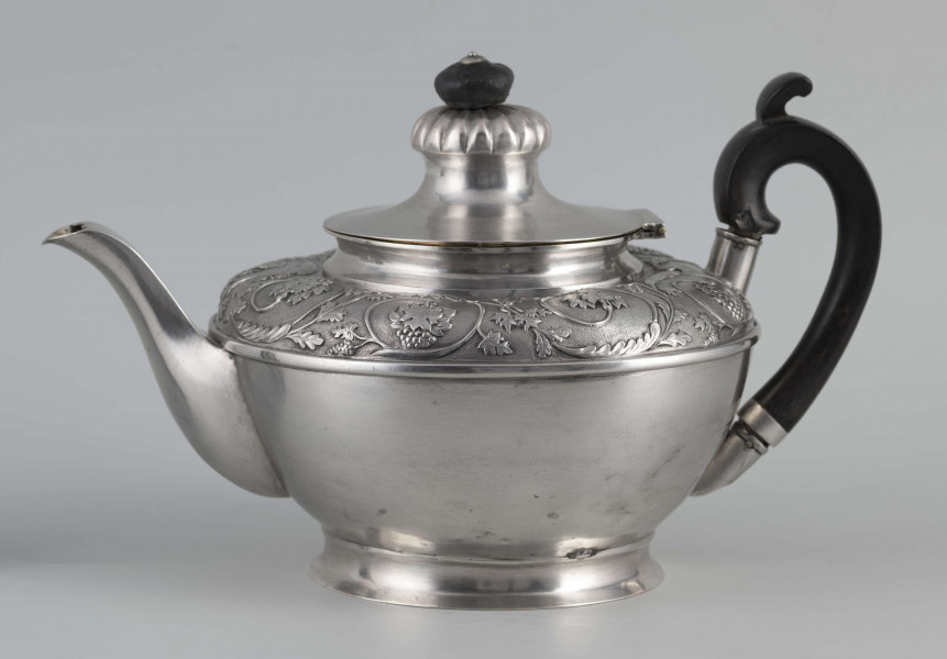 S/Mt/542/ML - Korpus w kształcie spłaszczonej kuli, na niskiej stopie, wylew w formie dzióbka, ucho drewniane malowane na czarno, osadzone w dwóch tulejkach, pokrywka na zawiasie, płaska, w części środkowej wypiętrzona i ozdobiona puklowaną rozetą.Górna część brzuśca ozdobiona sztancowaną taśmą z wzorem spiralnie zwinietej wici roślinnej z kwiatami, uchwyt przykrywki zakończony drewnianą rozetką.  
