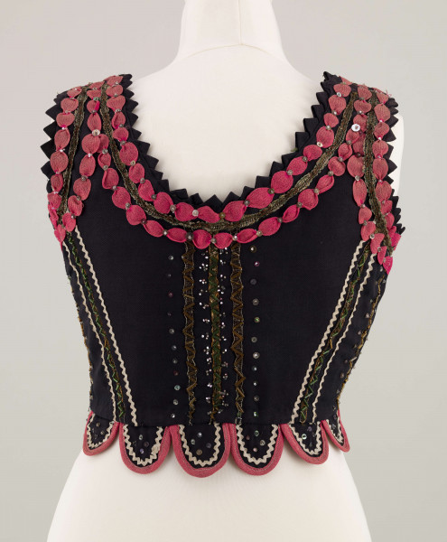 E/7358/ML - Gorset zszyty z 3 kawałków granatowej wełenki, podszyty brązową bawełnianą podszewką. Z przodu rozcięcie usztywnione fiszbinami, zaopatrzone w 10 haftek drucianych do przewlekania wstążeczki. Od pasa gorsetu dookoła opadają klapki. Cały gorset bogato zdobiony amarantowymi tasiemkami, żółtym i srebrnym szychem, białą krepiną, barwnymi koralikami i cekinami. Przód gorsetu, brzegi wykroju i pachy ozdobione dodatkowo granatowymi ząbkami. 