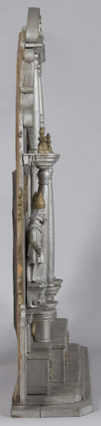 E/7373/ML - Ołtarzyk składa się z 3 części: podstawy i 2 pięter. Oba piętra podzielone na 3 pola (środkowe 24 cm, boczne 13,5 cm) przez kolumny. Podstawa posiadająca wysuniętą część środkową, ozdobiona jest 4 krzyżami greckimi wpisanymi w gwiazdę złot. Podstawę flankują 2 małe ołtarzyki: prawe brak przedstawienia, po lewej Madonna – złota postać na owalnym srebrnym polu, z granatową aureolą, nad nią na granatowym tle – 4 złote gwiazdki. Prawy ołtarzyk nad brakującym przedstawieniem ma złotą gwiazdkę. Lewemu jej brak. Górne pole podstawy zdobią 2 kolumienki oraz złote guzy pomiędzy. 
I piętro w części środkowej, pomiędzy kolumnami z tympanonem z głową aniołka i klęczącymi aniołami na daszku, postać Chrystusa Ukrzyżowanego w kolorze srebrnym, wraz ze złotymi  inskrypcją i pionowymi ząbkami. Po bokach, symetrycznie, przedstawienia  św. Mikołaja w złotej czapce biskupiej, na owalnej podstawie, z jedną ręką uniesioną , a druga opuszczoną trzymającą rękojeść. Ponad stylizowany złoty ornament roślinny. Całość piętra flankowana ornamentem przypominającym liście akantu i ślimacznice. 
II piętro w częściach bocznych zamknięte jest spływami wolutowymi , na polach po złotej kolumience/sterczynie i gwieździe. W części środkowej oleodruk ze św. Józefem z Dzieciątkiem, w złotej ramce. Całość wieńczą postaci ornatów w kolorze srebrnym i złote niepełne spływy wolutowe ze ślimacznicą. 
Brak wielu elementów. Plecy ołtarzyka stanowi 6 deseczek o szer. 10,5 cm i grubości 1,2 cm.