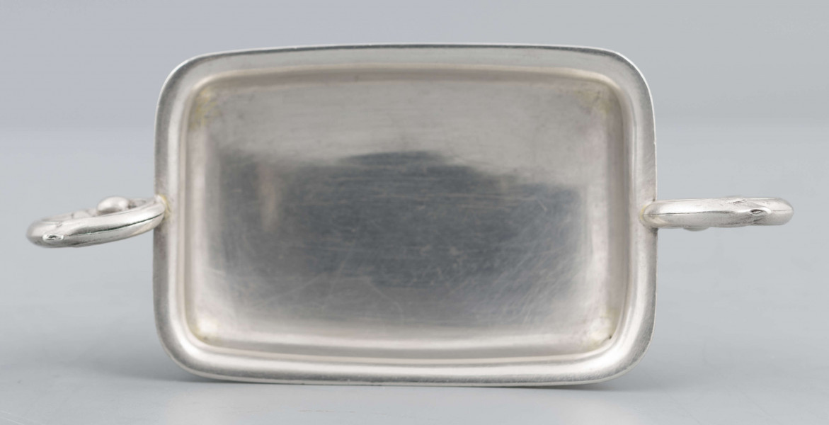 S/Mt/57/ML - Stopa prostokątna, wysklepiona, przechodząca w trzon z owalnym kapturkiem sztancowanym we wzór akantu. Czarka prostokatna o zaokrąglonych narożach i wywinietym brzegu, z dwoma uchami na krótszych bokach. Ucha wolutowe zdobione listkami. 