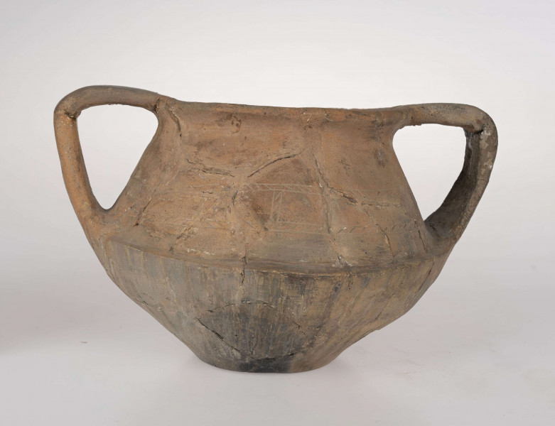 Naczynie wazowate, dwustożkowe z dwoma taśmowatymi uchami. Nad załomem brzuśca zdobienie w postaci linii rytych wypełnionych ukośnym kreskowaniem. Powierzchnia brunatna. Typ III/3 wg T. Liany.