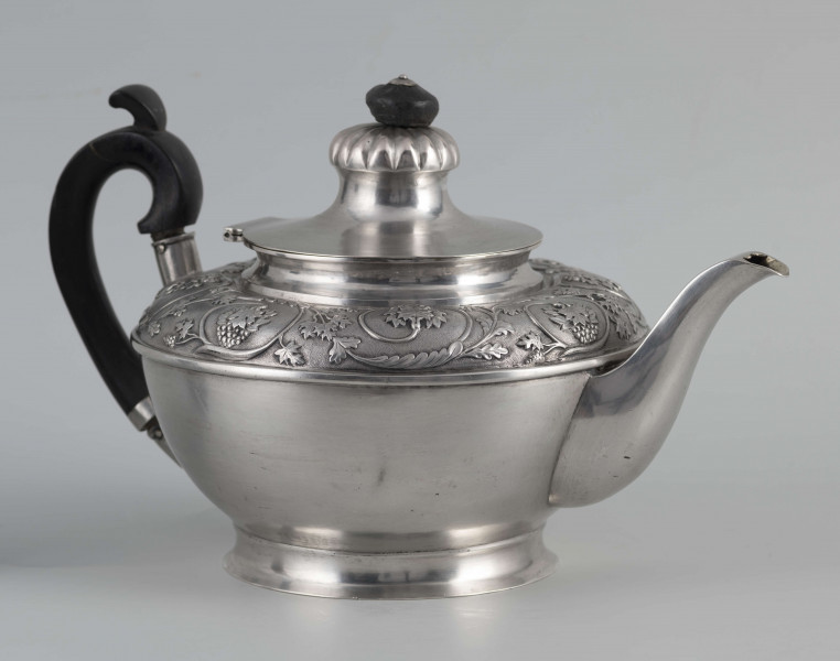 S/Mt/542/ML - Korpus w kształcie spłaszczonej kuli, na niskiej stopie, wylew w formie dzióbka, ucho drewniane malowane na czarno, osadzone w dwóch tulejkach, pokrywka na zawiasie, płaska, w części środkowej wypiętrzona i ozdobiona puklowaną rozetą.Górna część brzuśca ozdobiona sztancowaną taśmą z wzorem spiralnie zwinietej wici roślinnej z kwiatami, uchwyt przykrywki zakończony drewnianą rozetką.  
