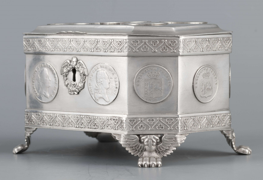 S/Mt/364/ML - Cukiernica skrzynkowa prostokątna ze ściętymi narożami, na czterech nóżkach w formie uskrzydlonych lwich łap, z wieczkiem o wklęsło profilowanym brzegu. Górny i dolny brzeg obiega  moletowana taśma z motywem neogotyckich trójliści, szyld zamka ma formę różanego wieńca pod skrzydatą główką..W ścianki i wieczko wmontowanych jest 15 monet srebrnych:
1 ort Zygmunta III
1 talar pruski, 1818 r.
1 pięciofrankówka, 1811 r.
1 moneta austriacka, 1797 r.
1 moneta saska, 
2 monety polskie St.Augusta Poniatowskiego
1 dwózłotówka Królestwa Polskiego, 1825 r.
1 pięciozłotówka, 1831 r.
6 złotówek, 1831 r.
Na dnie sygnatura wklęsła kursywą:
- K. Rotkiel w Lublinie
i punca z oznaczeniem próby 