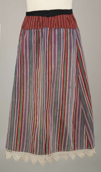 E/8132/ML - Fartuch wełniany szyty maszynowo. Wykonany z materiału samodziałowego tkanego w pionowe paski, w kolorach: czerwonym, białym, szafirowym, bordowym, zielonym. Uszyta z prostokątnego kawałka materiału, do którego doszyto z obu stron kliny. U góry fartuszek przedłużony przez wszycie kawałka materiału szer. 7 cm, w pasy czerwono-granatowe. W talii wszyty w pasek z czarnego materiału, szer. 2 cm, zakończony troczkami. Dół fartuszka zdobi biała koronka szer. 5 cm.